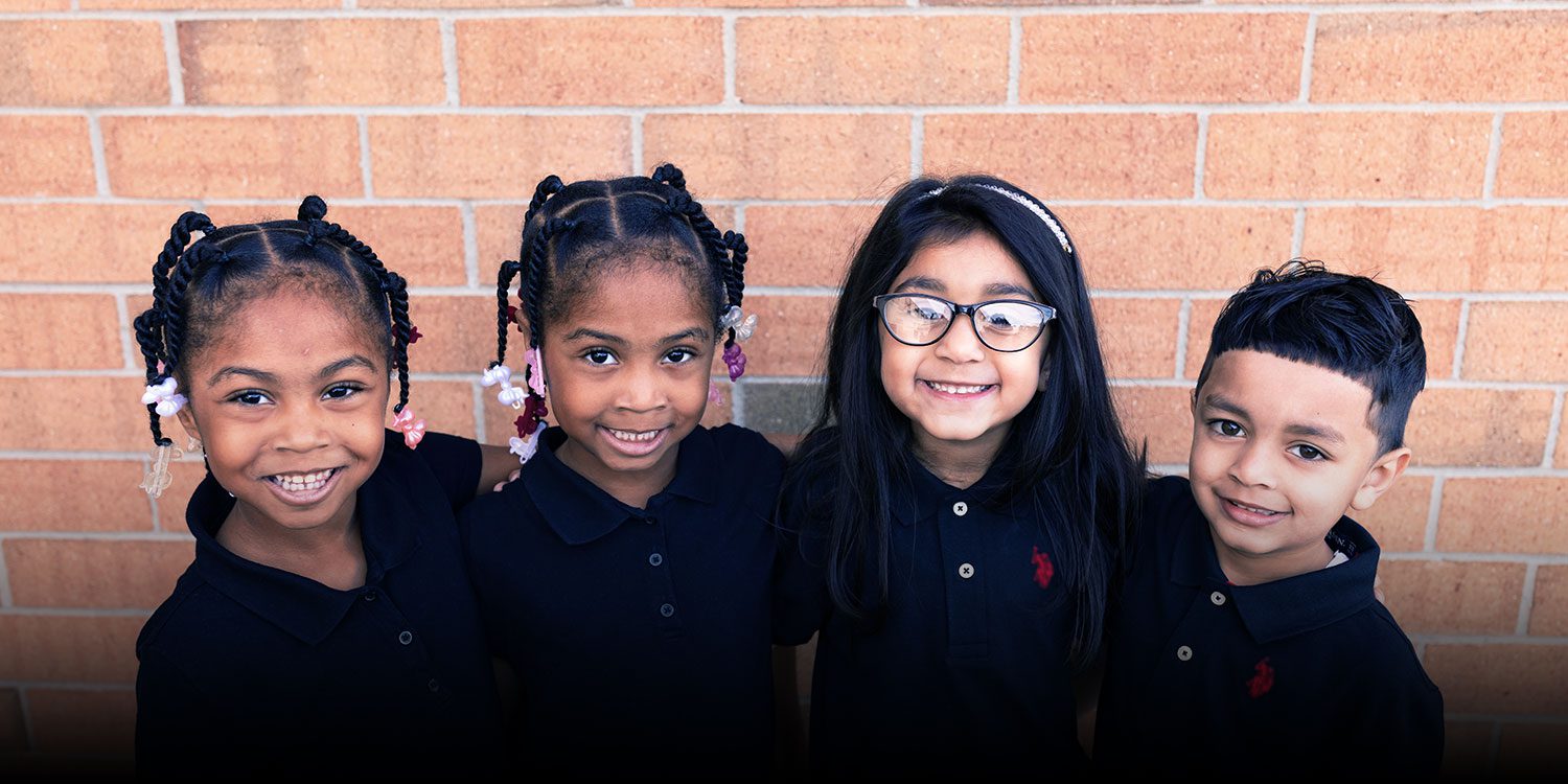 Four smiling students standing together against a brick wall.