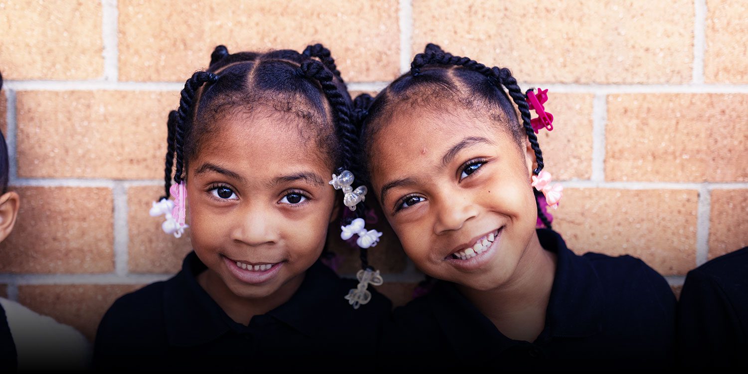 Two smiling students against a brick wall.